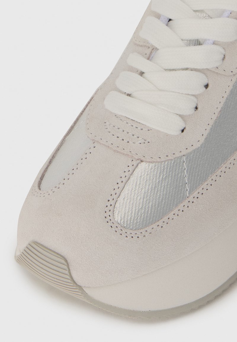 Sneaker in suede grigio chiaro con accenti metallici argento, punta arrotondata e suola testurizzata. Presenta lacci bianchi e decorazioni a cucitura.