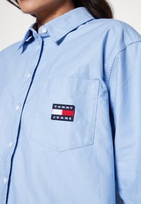 Light blue button-up shirt in cotton with a chest pocket, featuring a rectangular logo patch in navy, red, and white, with the text "TOMMY JEANS."