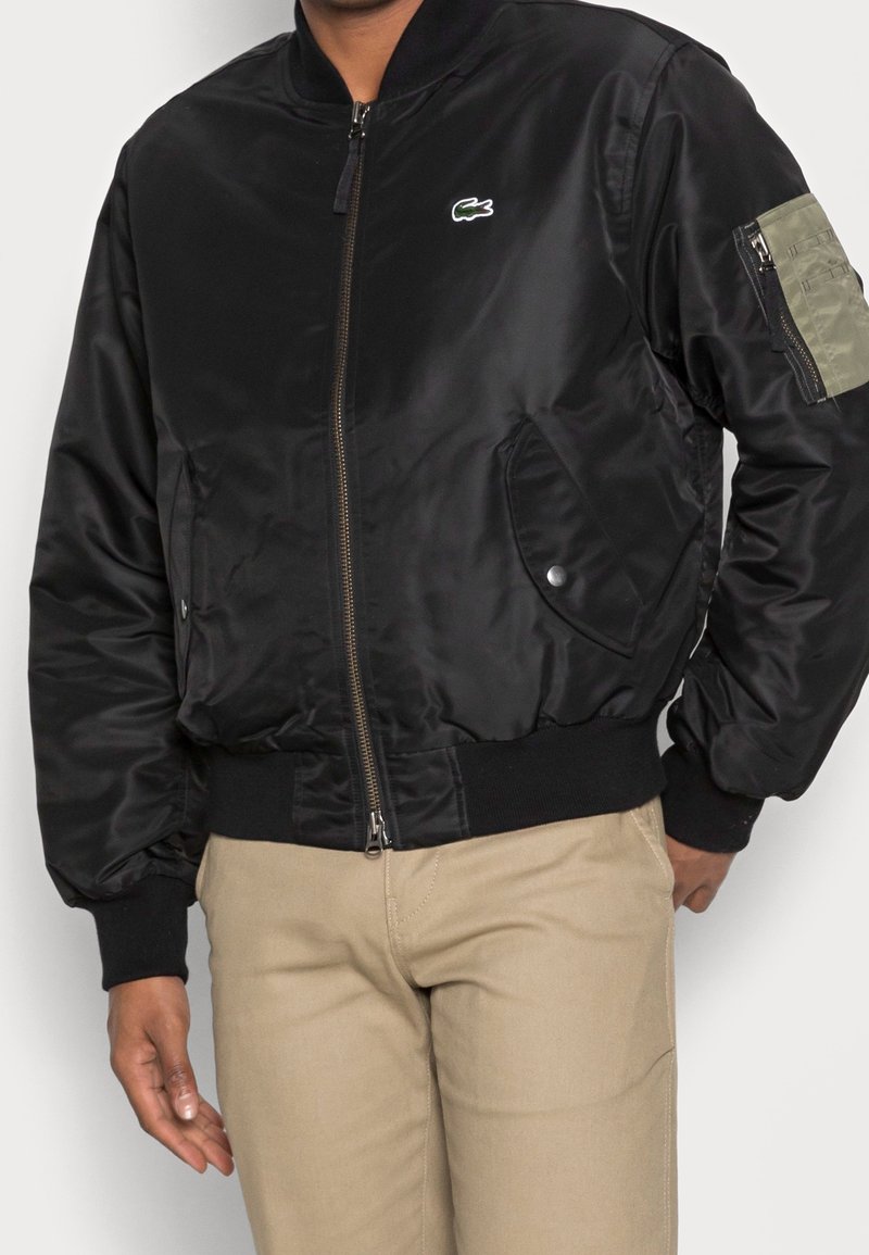 Homme portant une veste bomber noire zippée avec un petit logo crocodile vert sur la poitrine, associée à un pantalon beige.