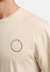 Camisa de manga larga beige fabricada en suave algodón. Presenta un diseño impreso de texto redondo en verde oliva en el pecho. Cuello redondo clásico.