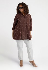 Camicia marrone con bottoni, decorata con un motivo geometrico, maniche a tre quarti, abbinata a pantaloni bianchi e tacchi color nude. Occhiali da sole trasparenti indossati.