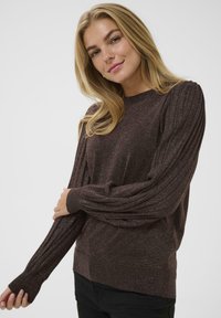 Blondide juustega naine, kes kannab pikka varrukat, tumepruunis helkiva kootud sviitrit, poseerides üks käsi üle keha.