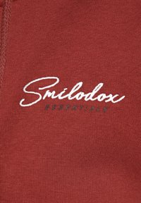 Tkanina w kolorze rdzy o gładkiej fakturze, na której białą czcionką napisano "Smilodox", a poniżej w ciemnozielonym "ESSENTIALS".