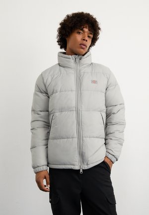 Chaqueta acolchada gris claro con alto cuello, cierre con cremallera, secciones cosidas y un pequeño parche de logo en el pecho. Textura suave y forrada.