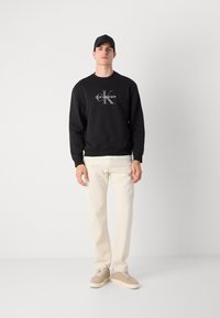 Sweatshirt en coton noir avec le logo "Calvin Klein" blanc, associé à un jean de couleur claire et des baskets beiges. Design simple avec des bords côtelés.