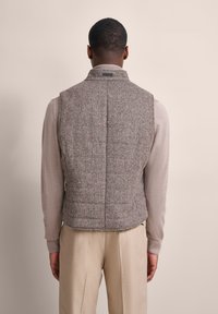 Brauner Hahnentrittmuster-Pufferweste mit hohem Kragen, gestepptem Design, Schlitz im Rücken und Logo-Detail am Ausschnitt, getragen über einem hellen Pullover.