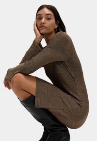 Bronze schimmerndes Langarmkleid mit figurbetontem Schnitt und seitlichem Schlitz, kombiniert mit kniehohen schwarzen Stiefeln. Glatte, reflektierende Textur.