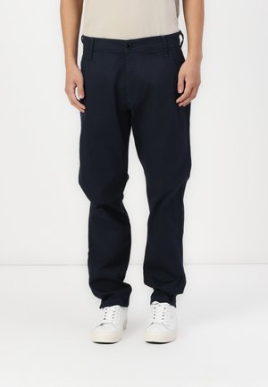 Vue de face d'une personne portant un pantalon bleu marine foncé, une chemise beige et des baskets blanches, debout devant un fond blanc uni.
