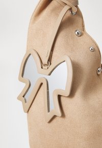 Beige semsket veske med en bemerkelsesverdig sløyfeformet speilaccessoire. Satinaktig tekstur med sølvfargede detaljer og et mykt, buet design.