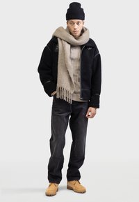 Schwarze Fleecejacke mit Reißverschluss, beiger gestrickter Schal, hellfarbiges Hemd mit Knöpfen, dunkle Jeans und beige Schuhe. Akzente umfassen kontrastierende Texturen.
