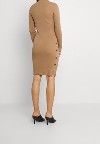 Robe midi en maille beige avec une silhouette ajustée. Elle présente un col montant, des manches longues, des boutons latéraux et une fente à l'ourlet. Mules à talons noires.