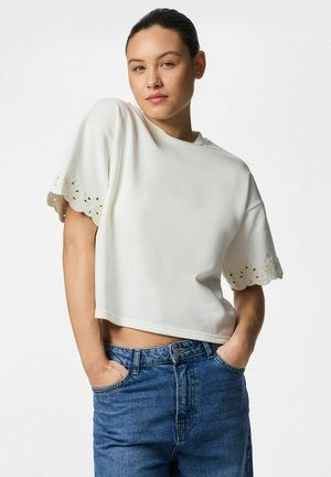 Nuori nainen tummatukka takana sidottuna, valkoinen lyhythihainen pusero jossa rei'ityskoristeita ja siniset korkeavyötäröiset farkut, kädet taskuissa.