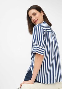 Camicia a righe navy e bianca, maniche corte con dettagli a bottoni, vestibilità comoda, tessuto liscio con piega sul retro per facilitare i movimenti.