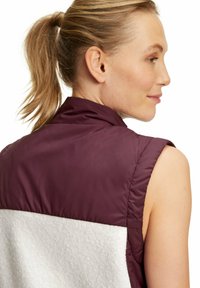 Ärmellose Weste mit einem tiefburgunderfarbenen oberen Bereich und einem weißen, strukturierten Fleece-Unterteil, mit sichtbaren Nähten und taillierter Passform.