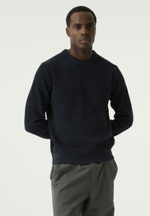 Man standing against a plain background, wearing a dark ribbed sweater and gray pants, facing forward with hands behind his back.