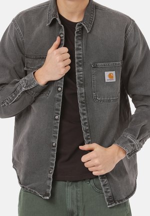 Person, der lukker en grå denimskjorte med knapper over en sort T-shirt og grønne bukser, og viser Carhartt-logoet på venstre brystlomme.