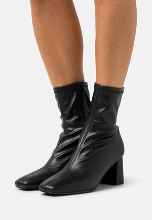 Botas de tobillo negras hechas de cuero suave, con un diseño liso y caído, punta afilada y un tacón cuadrado para mayor estabilidad.