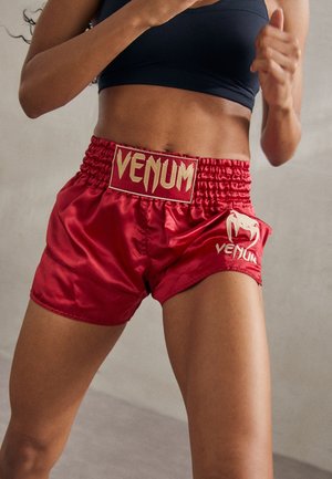 Atleta con pantaloncini rossi Venum e top sportivo nero in posizione da boxe con i pugni alzati a metà movimento.