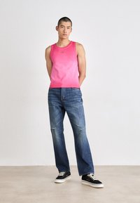 Pink gradient tank top, blue denim jeans with distressed accents, black sneakers with white soles, standing on a light beige floor.