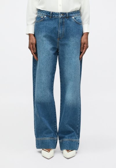 Jean large bleu clair en denim avec taille haute, dotés de poches avant et de détails de couture contrastants. Portés avec des chaussures blanches.