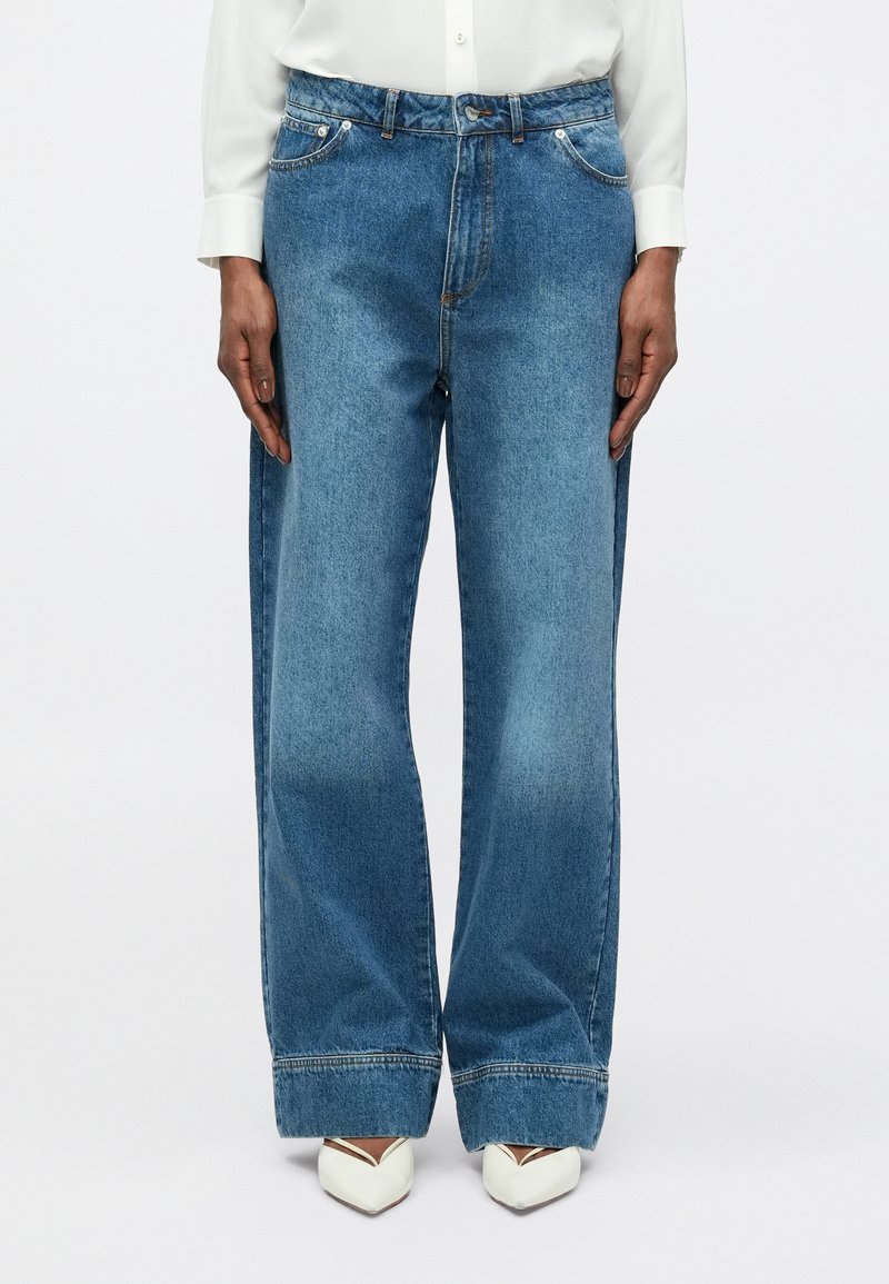 Jeansi larga, de culoare albastru deschis, fabricați din denim, cu talie înaltă, având buzunare frontale și detalii de cusătură în contrast. Asortați cu încălțăminte albă.