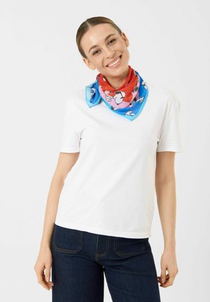 Femme souriante portant un t-shirt blanc, un jean foncé et un foulard coloré imprimé Snoopy autour du cou sur un fond uni.