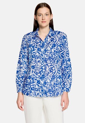 Vrouw draagt een wit overhemd met blauwe bloemenpatronen en witte broek, staand tegen een effen achtergrond.