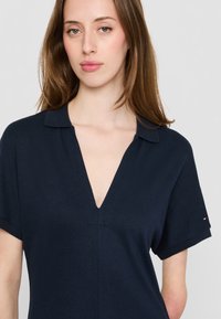 Marineblå polo skjorte med dyp V-hals og korte ermer. Laget av teksturert stoff, med diskrete sømmer og en liten logo på ermet.