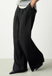 Pantaloni negri cu croială largă dintr-un material ușor cu o textură netedă. Prezintă buzunare laterale și o croială lejeră, asortați cu pantofi negri cu vârf ascuțit.