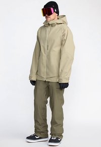 Veste imperméable beige avec capuche, associée à un pantalon vert olive. Comprend des gants noirs et des lunettes de protection miroir surdimensionnées. Chaussures noires avec des accents blancs.