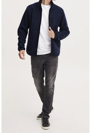 Man draagt een marineblauwe fleecejas met rits over een wit T-shirt, versleten zwarte spijkerbroek en zwarte sneakers, lopend naar voren op een lichte achtergrond.