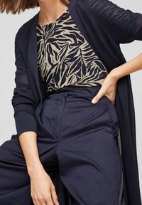 Cardigan de punto abierto en color azul marino sobre una blusa beige con rayas de cebra negras, combinado con pantalones de tiro alto en azul marino. Enfócate en los detalles de textura y patrón.