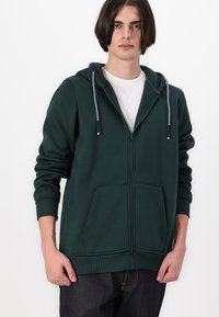 Zöld, cipzáros kapucnis pulóver, puha anyagból készült, kapucnival, két elülső zsebbel és kontrasztos húzókábellel. Fehér póló fölé van viselve.