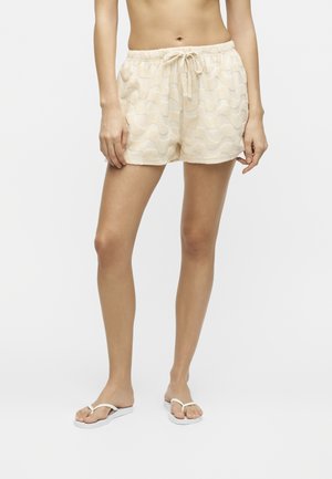 Γυναίκα που φορά μπεζ σορτς με σχέδιο και κορδόνι και λευκά σαγιονάρες, στέκεται μπροστά σε ένα μονόχρωμο φωτεινό φόντο.