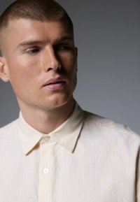 Cream button-up shirt with thin yellow stripes, featuring a pointed collar and textured fabric, partially buttoned at the neck.