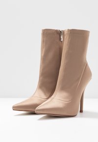 Botines beige de punta afilada con tacones de aguja altos y cremalleras laterales, exhibidos sobre una superficie blanca con un fondo sencillo.