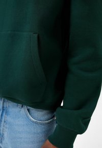 Felpa verde con tasca frontale, polsini e bordo a costine. Il materiale sembra morbido; abbinata a jeans in denim azzurro chiaro.