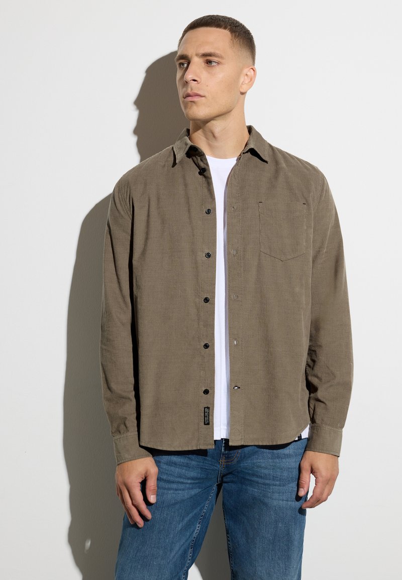 Brown button-up shirt with a textured finish, single chest pocket, worn over a white t-shirt, paired with blue jeans.