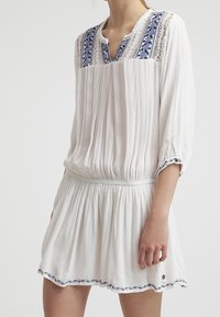 Vit klänning med en texturerad tyg, med blå broderade detaljer vid halsringningen och ärmarna, samt en rynkad midja för en utsvängd kjol.