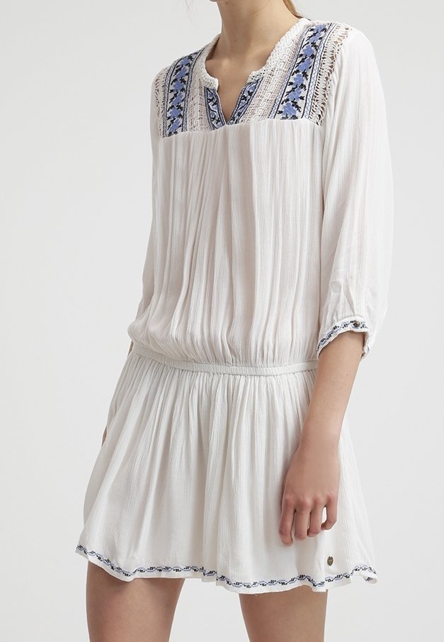 Vit klänning med en texturerad tyg, med blå broderade detaljer vid halsringningen och ärmarna, samt en rynkad midja för en utsvängd kjol.
