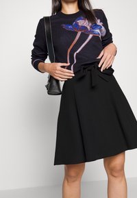 Jupe noire avec un nœud à la taille, associée à un pull sombre avec un motif floral en bleu et rose. Matériaux lisses et texturés.