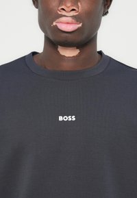 Camiseta de gola redonda cinza-escura, feita de um tecido suave, apresentando um logotipo branco "BOSS" no peito. Textura suave, corte clássico.