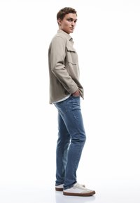 Beżowa koszula na guziki z kieszenią, jasnoniebieskie dżinsy i białe trampki z brązowymi podeszwami. Materiał wydaje się gładki i codzienny.