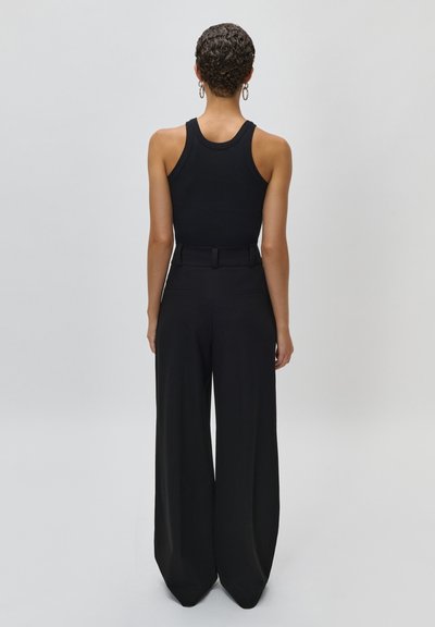 Frau mit kurzen lockigen Haaren, die ein schwarzes ärmelloses Oberteil und schwarze Hosen mit weitem Bein trägt, steht mit dem Rücken zum Betrachter vor einem einfarbigen Hintergrund.