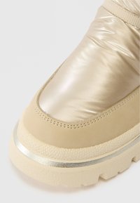 Beige boot with shiny padded fabric upper, suede trim, textured sole, and silver metallic accent on the edge of the sole.