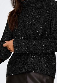 Femme portant un pull à col roulé noir scintillant à manches longues, associé à un pantalon en cuir foncé.