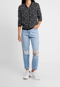 Veste bomber noire avec motif floral blanc, fermeture éclair, poignets côtelés, portée avec un jean bleu clair déchiré et des baskets blanches.