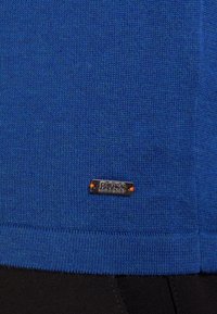 Blue knitted fabric with a small metal tag stitched with orange thread reading "BOSS Hugo Boss" near the hem above black fabric.
