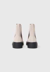 Et par beige semskede ankelstøvler med sorte chunky såler, set fra bagsiden på en ensfarvet hvid baggrund.