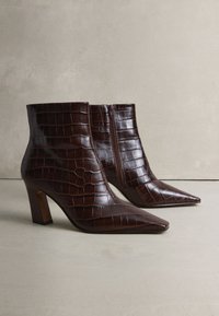 Dunkelbraune Stiefeletten aus strukturiertem Kunstkrokodilleder. Sie verfügen über eine spitze Zehenpartie, einen Blockabsatz und einen seitlichen Reißverschluss zum Schließen.